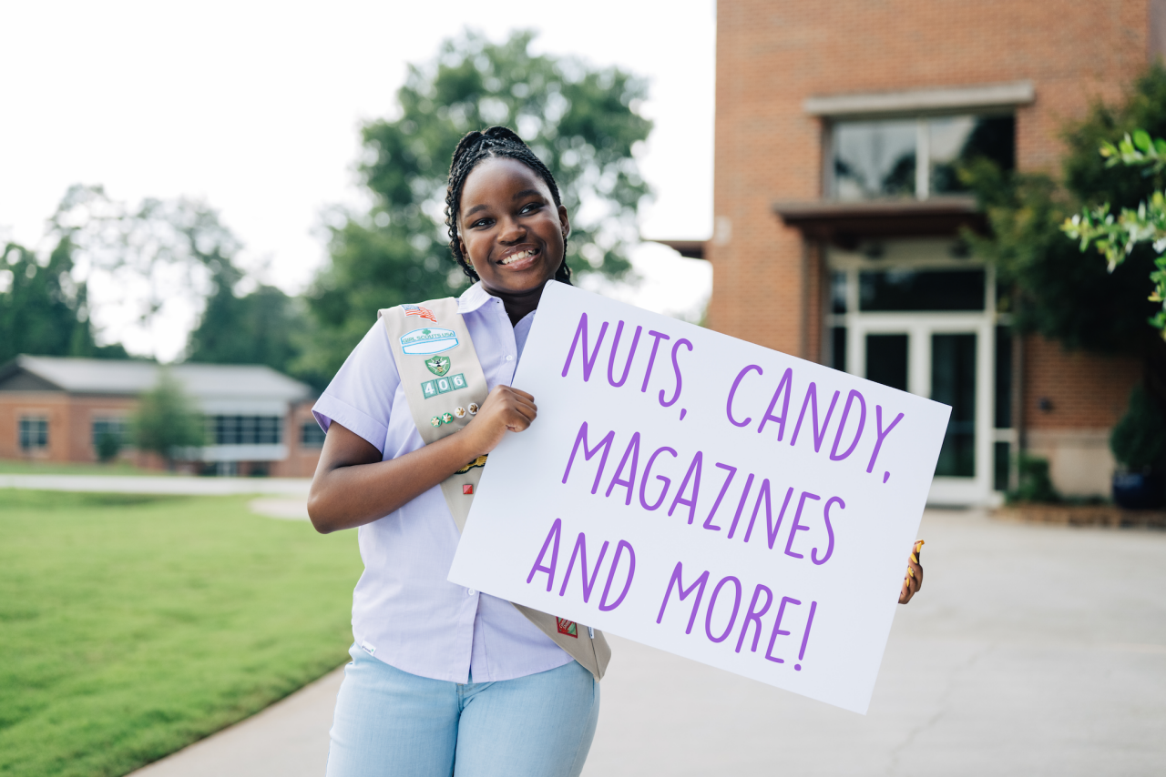 Cadette Girl holding a sign that reads "Nuts, Candy, Magazines, and More"!